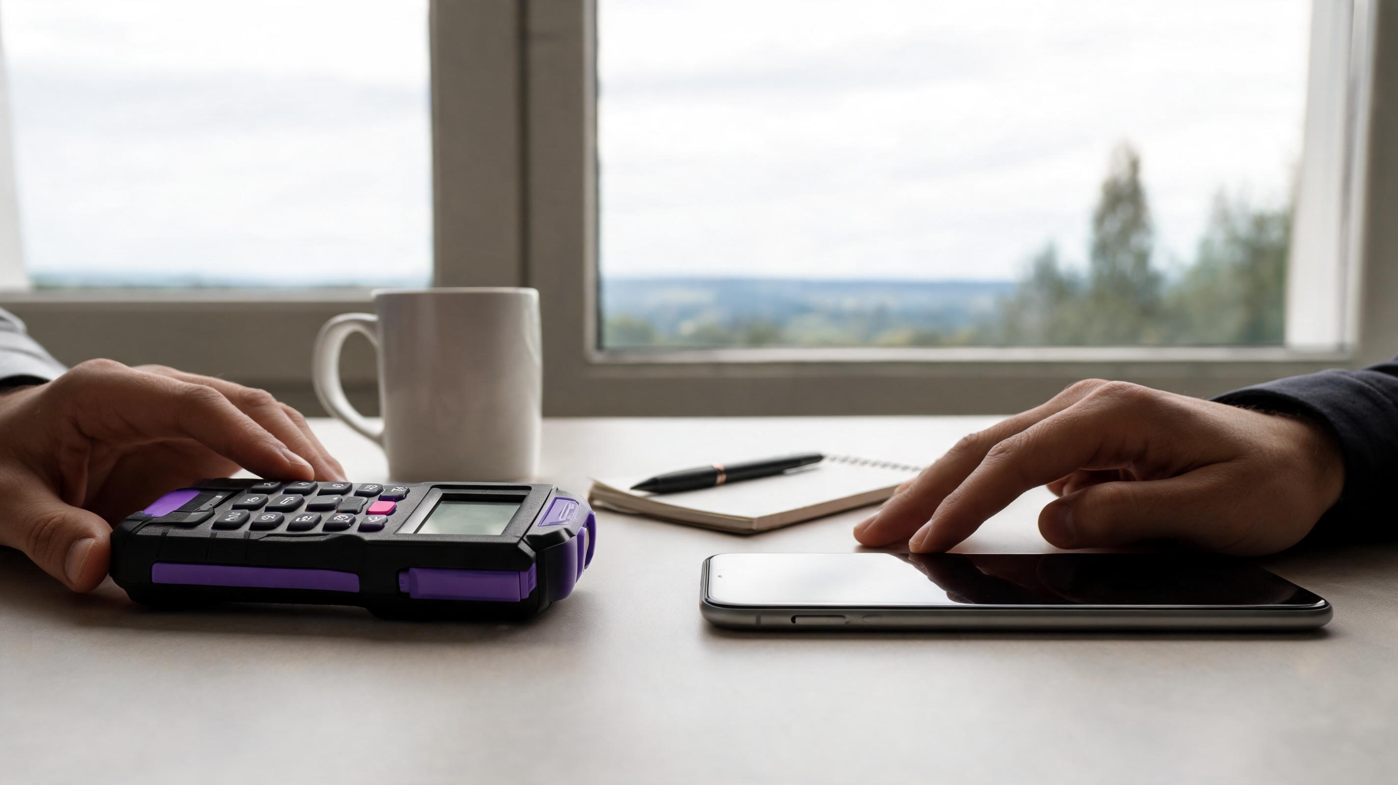 A close-up of two hands, one on a calculator and the other on a smartphone, with a coffee cup and notebook nearby